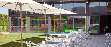 An outdoor patio area with tables and chairs, umbrellas, and a building with large windows and a tiled roof.