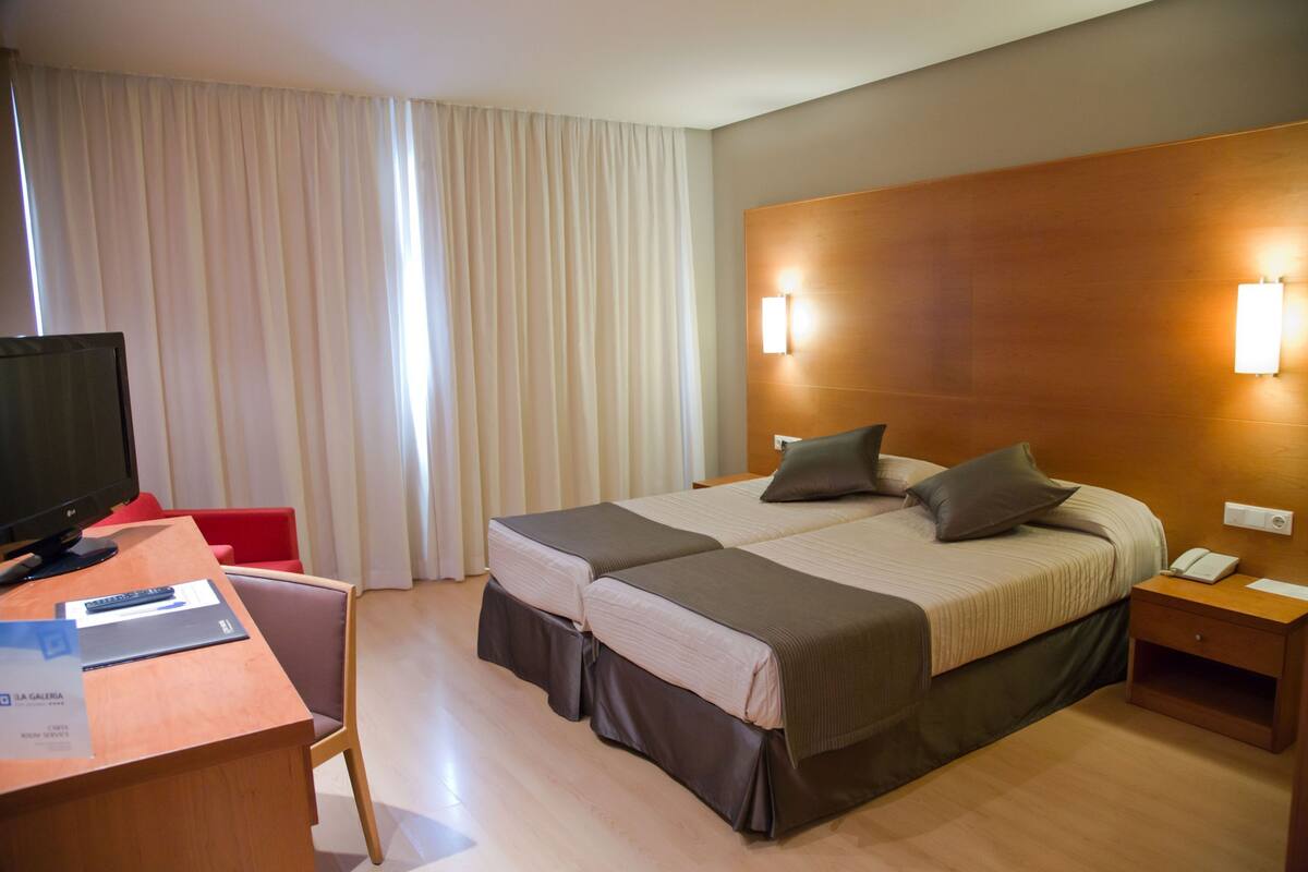 A hotel room with a bed, bedside tables, a desk with a computer, a TV, and curtains.