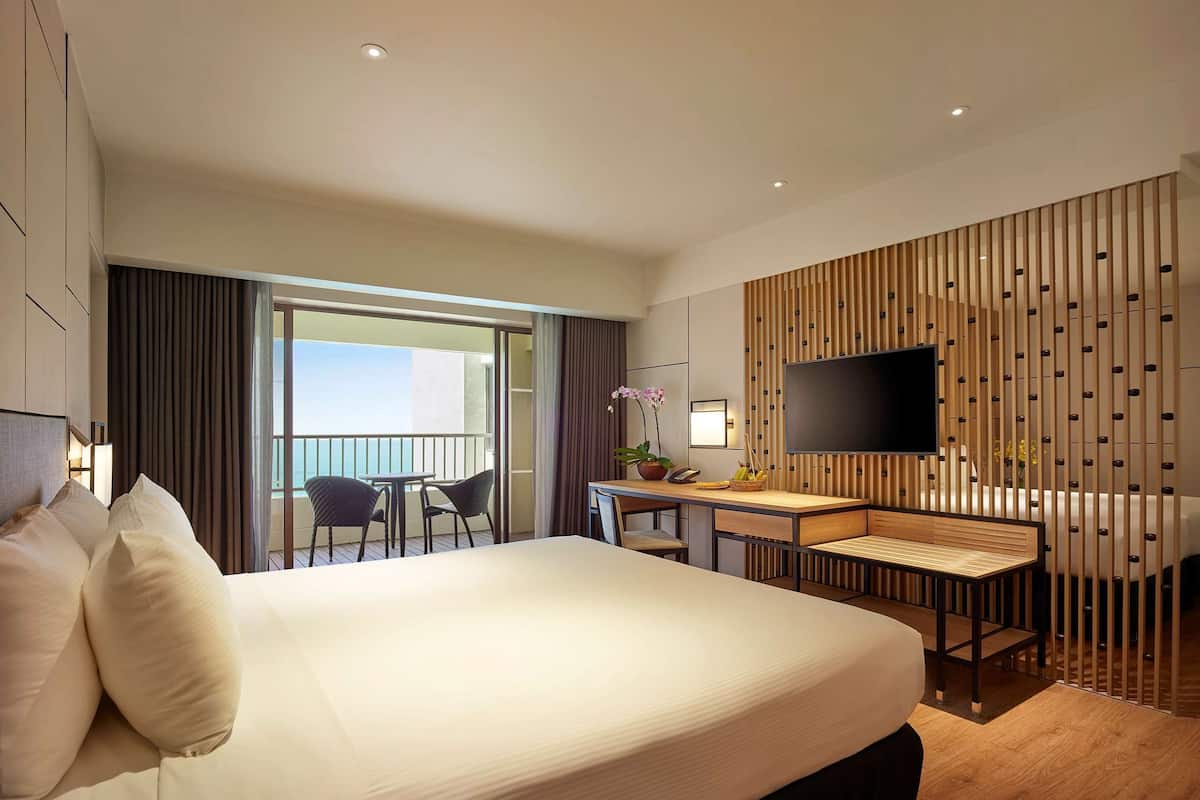 A hotel room with a large bed, a desk with a chair, a TV, and a balcony with a view.