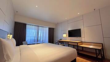 A hotel room with a large bed, a desk with a chair, a TV mounted on the wall, and a window with curtains.
