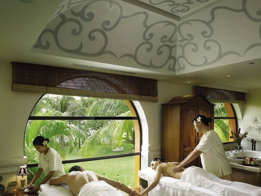 Un cuarto de spa con dos camillas de masaje, un ventanal con vistas a follaje y un techo con diseños decorativos.