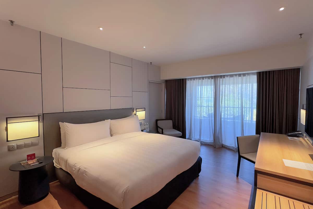 A hotel room with a large bed, bedside tables with lamps, a desk with a chair, and a large window with curtains.