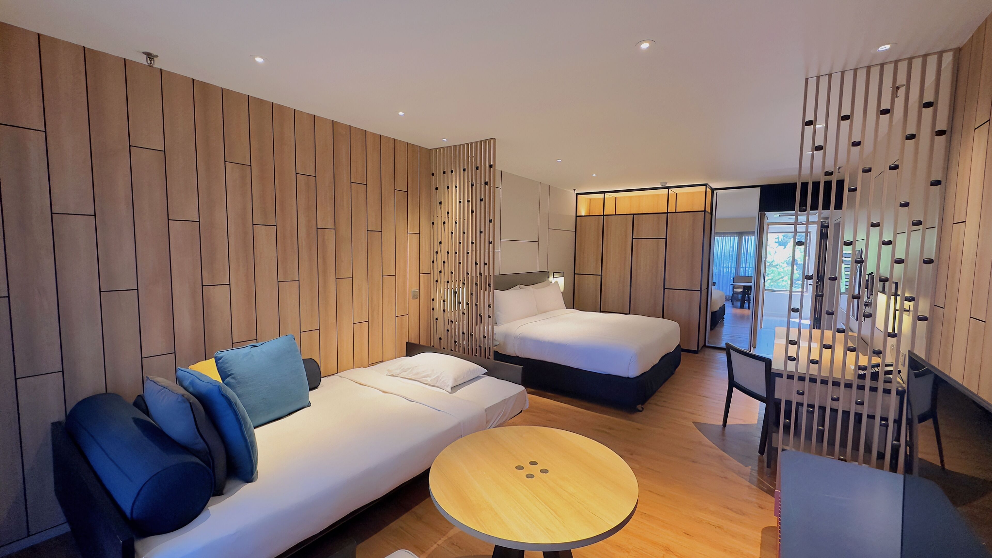 A modern hotel room with a wooden panel wall, a sofa with blue cushions, a bed, a round wooden table, and a balcony with a railing.