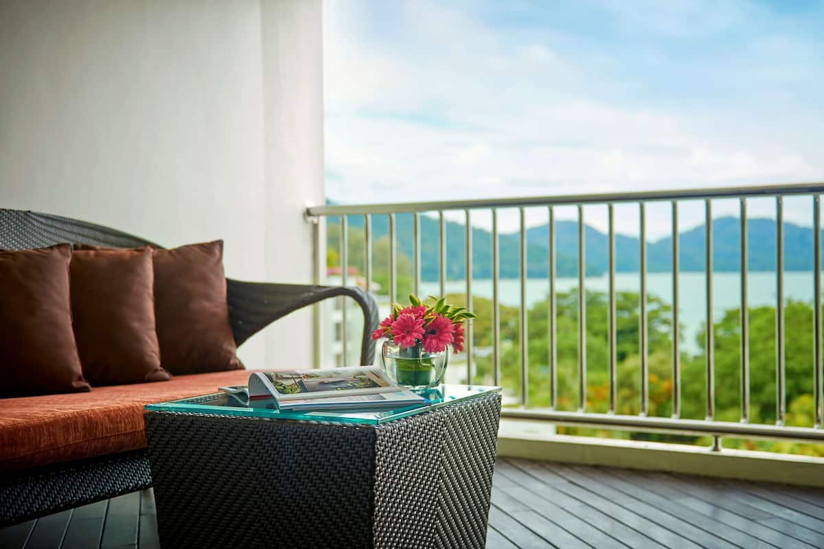 A balcony with a wicker sofa, a glass table with a vase of flowers, and a view of a lake and mountains.