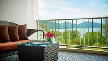 A balcony with a wicker sofa, a glass table with a vase of flowers, and a view of a lake and mountains.