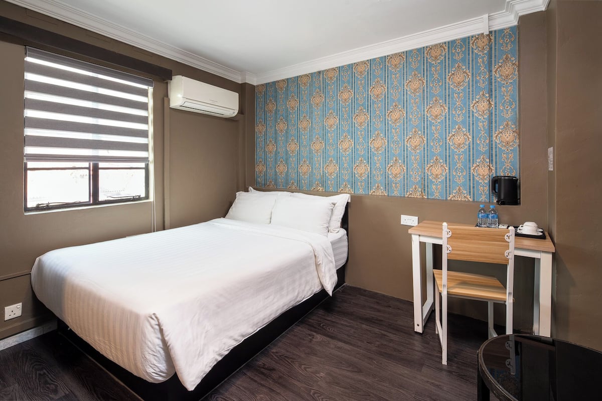 A bedroom with a bed, a small table with a chair, a window with blinds, and a patterned wall.