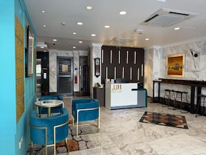 Un vestíbulo de hotel con un mostrador de recepción, sillones azules, piso de mármol y un reloj en la pared.