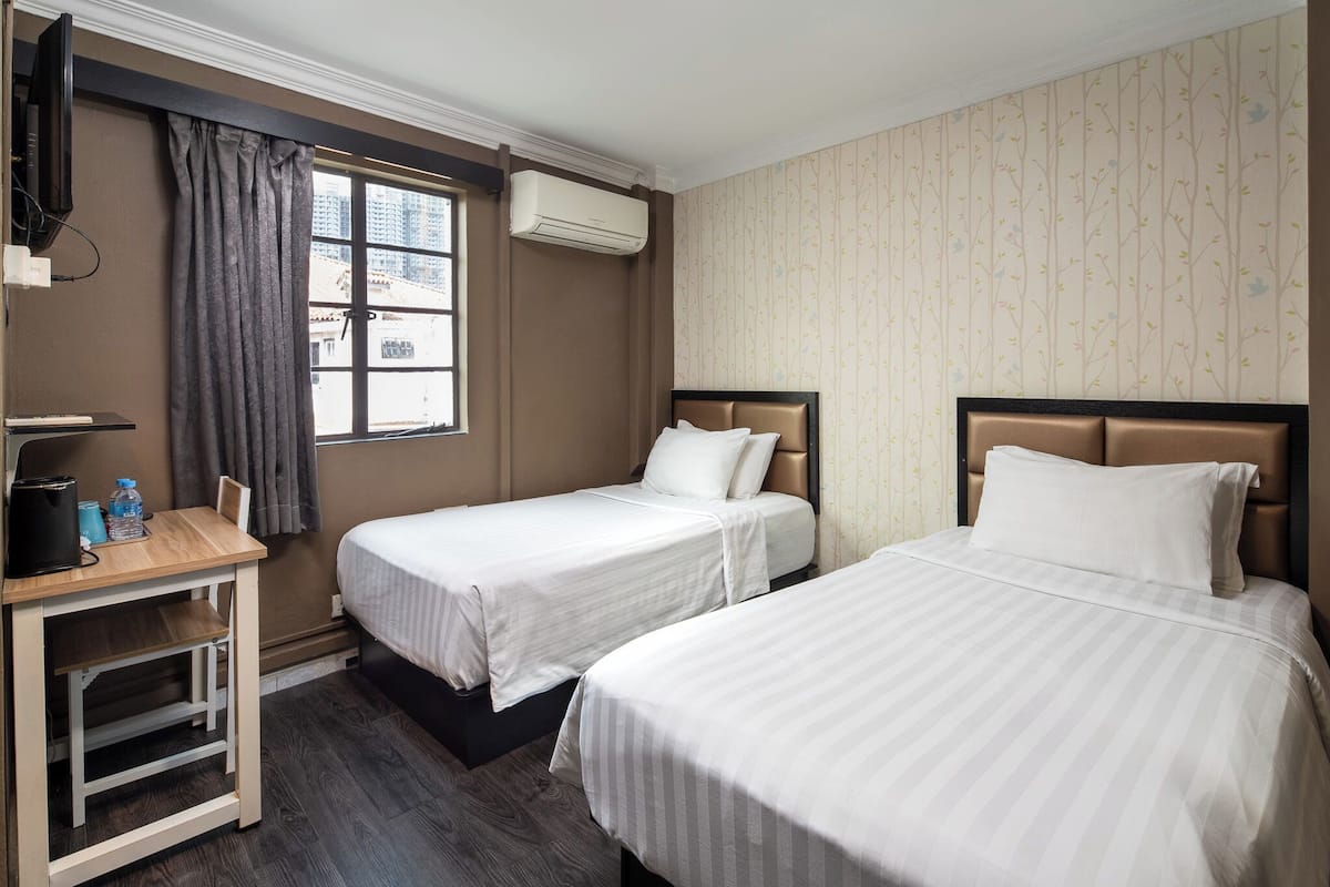 A hotel room with two single beds, a desk with a chair, a TV, and an air conditioning unit.