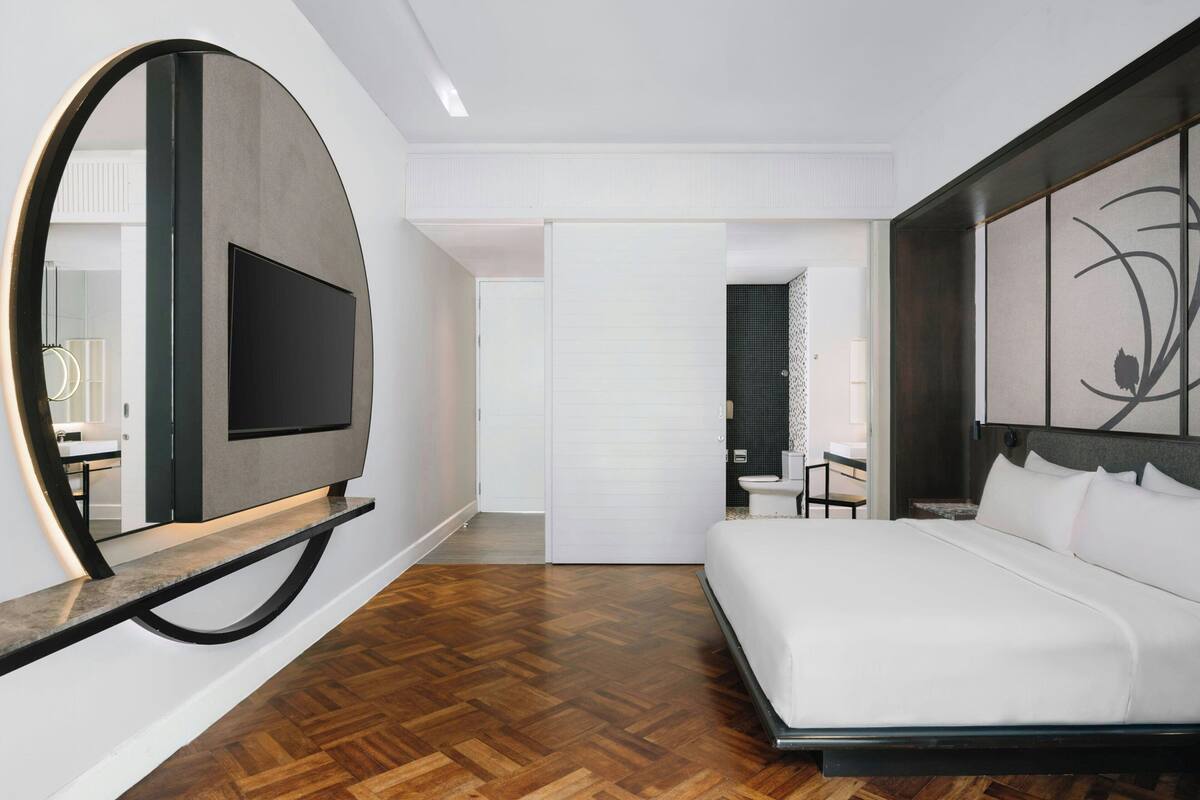 Une chambre d’hôtel moderne avec un grand lit, un téléviseur à écran plat fixé au mur, un miroir rond et un plancher en bois.