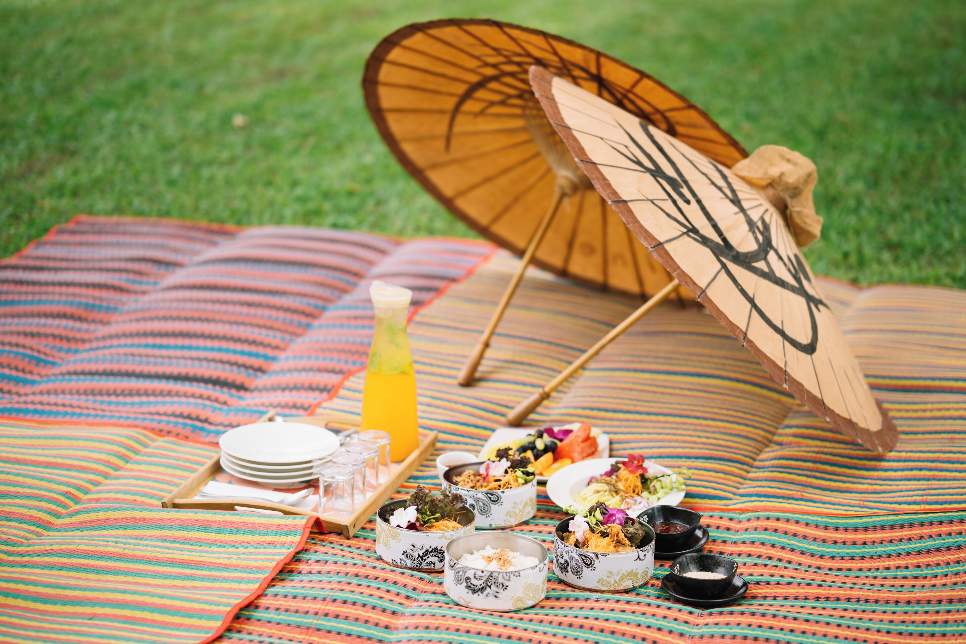 A picnic setup with a blanket, plates, bowls, and a wooden umbrella on a grassy area.