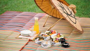A picnic setup with a blanket, plates, bowls, and a wooden umbrella on a grassy area.