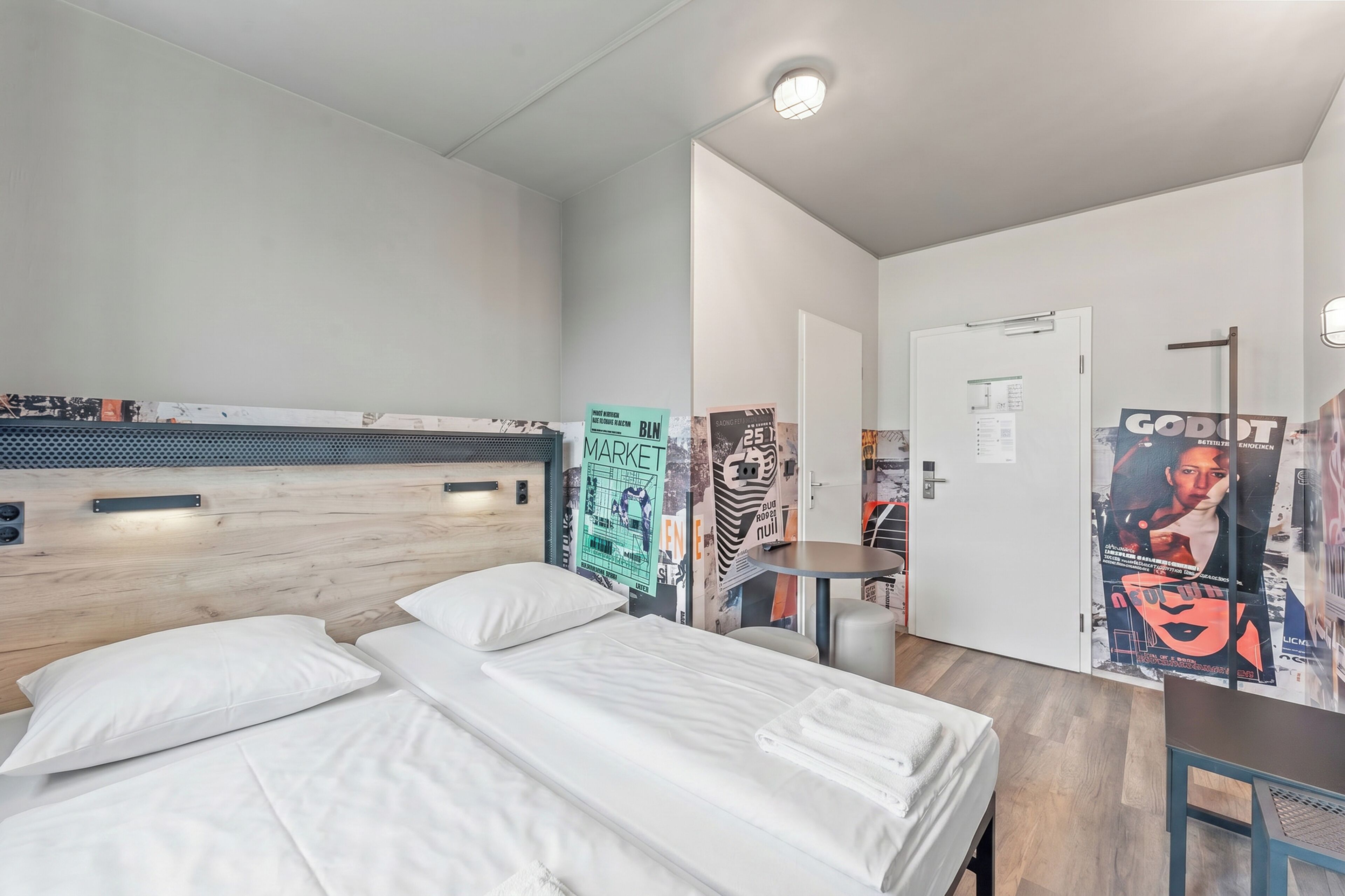 A modern hotel room with two beds, a small table, and a wall with posters.