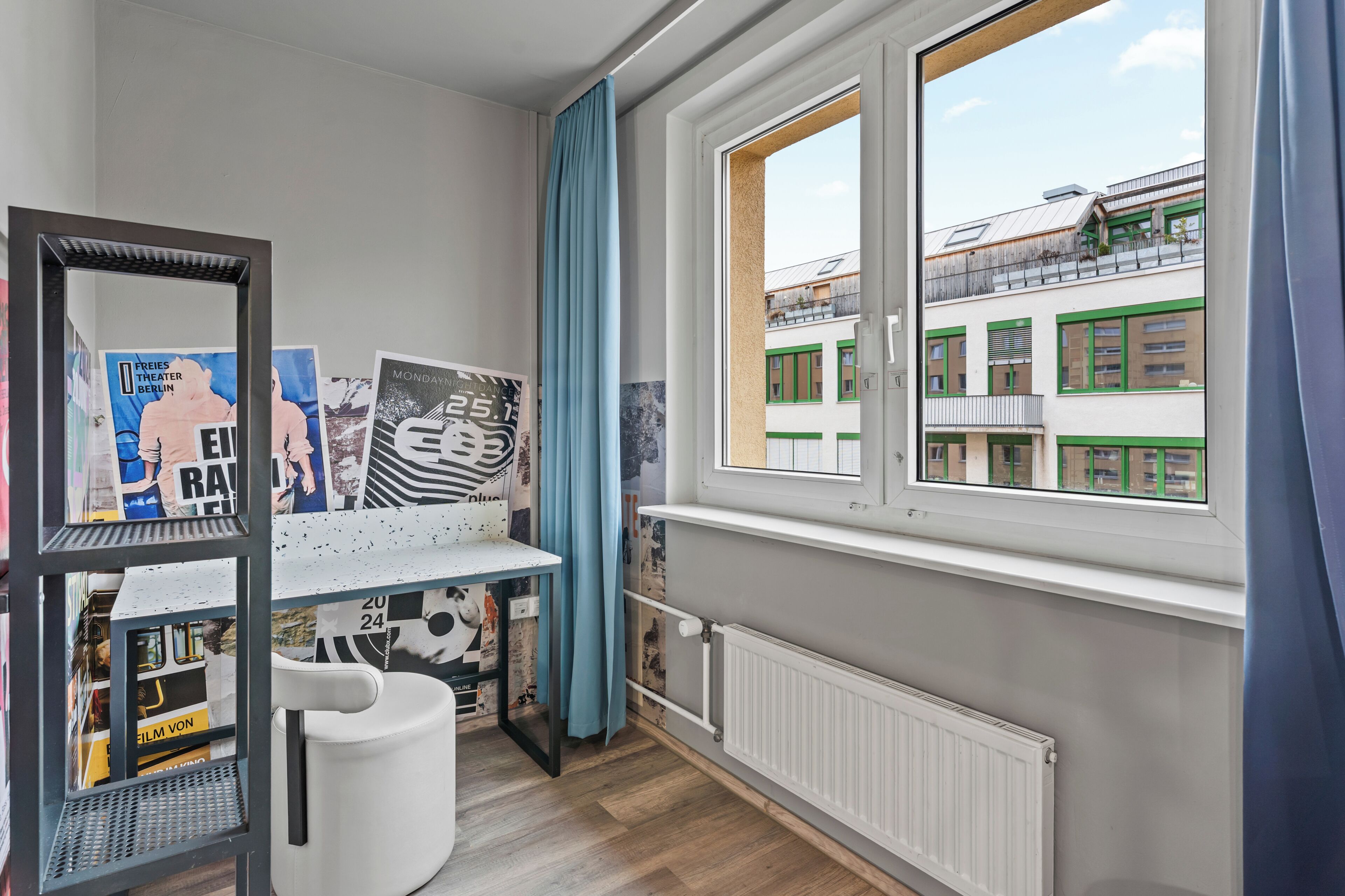 A room with a window, a radiator, a shelf with posters, and a chair.