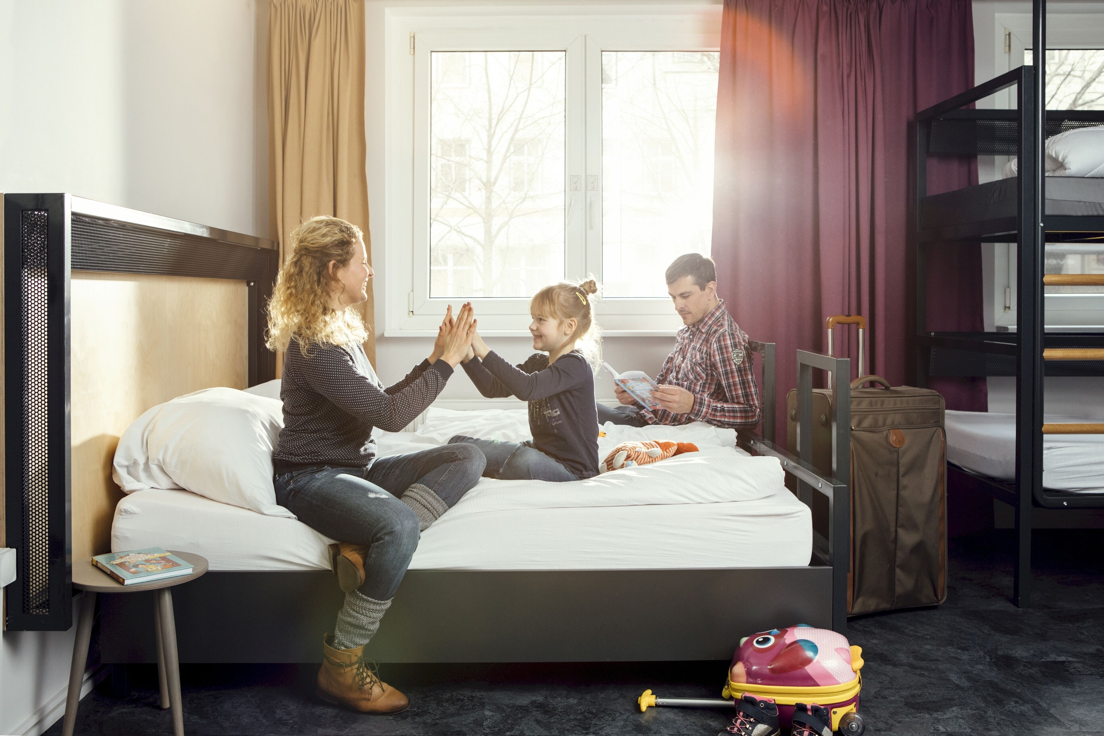 A hotel room with a bunk bed, a small bedside table, a window with curtains, and a toy on the floor.