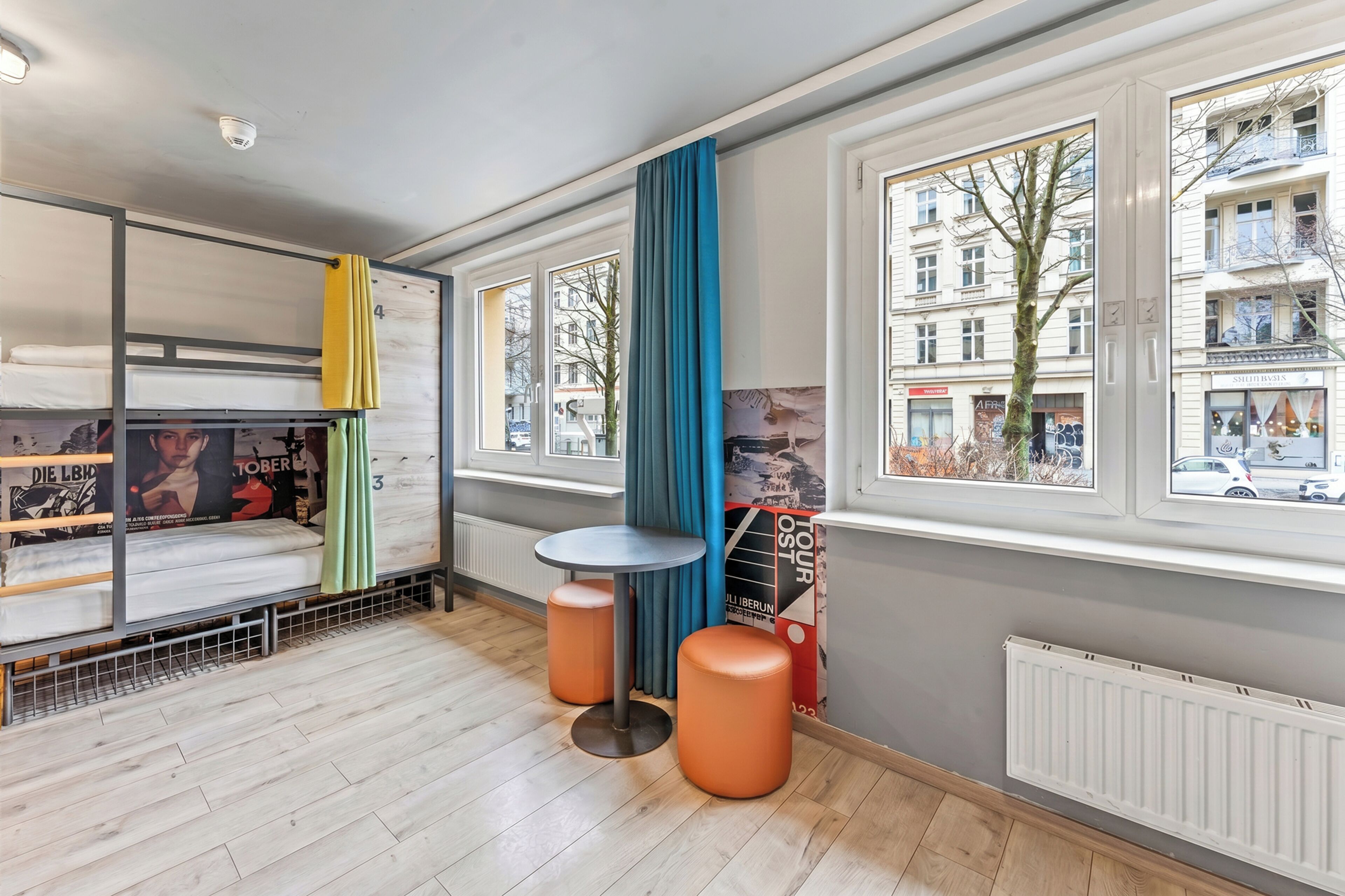 Ein moderner, gut beleuchteter Raum mit Etagenbetten, einem runden Tisch und Blick auf eine Stadtlandschaft durch große Fenster.