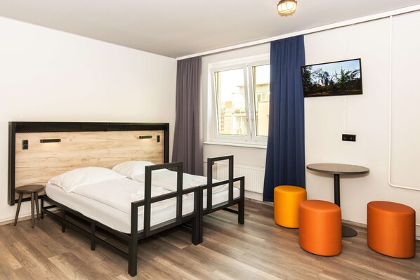 A modern bedroom with a wooden headboard bed, two orange ottomans, a small round table, and a window with curtains. - a&o Berlin Mitte (Berlin)