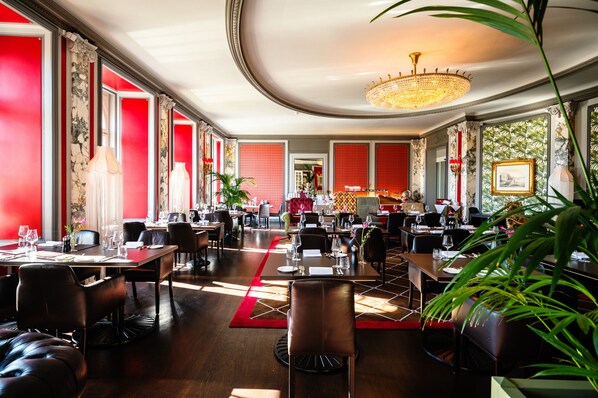 Un restaurant spacieux avec de hauts plafonds, des lustres et des touches de rouge. Les tables sont dressées avec des verres à vin et des menus.
