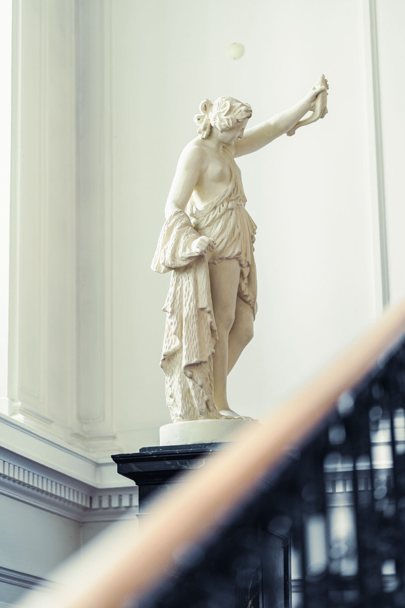 A marble statue of a woman in classical attire, standing on a pedestal in a well-lit room with white walls and a column.