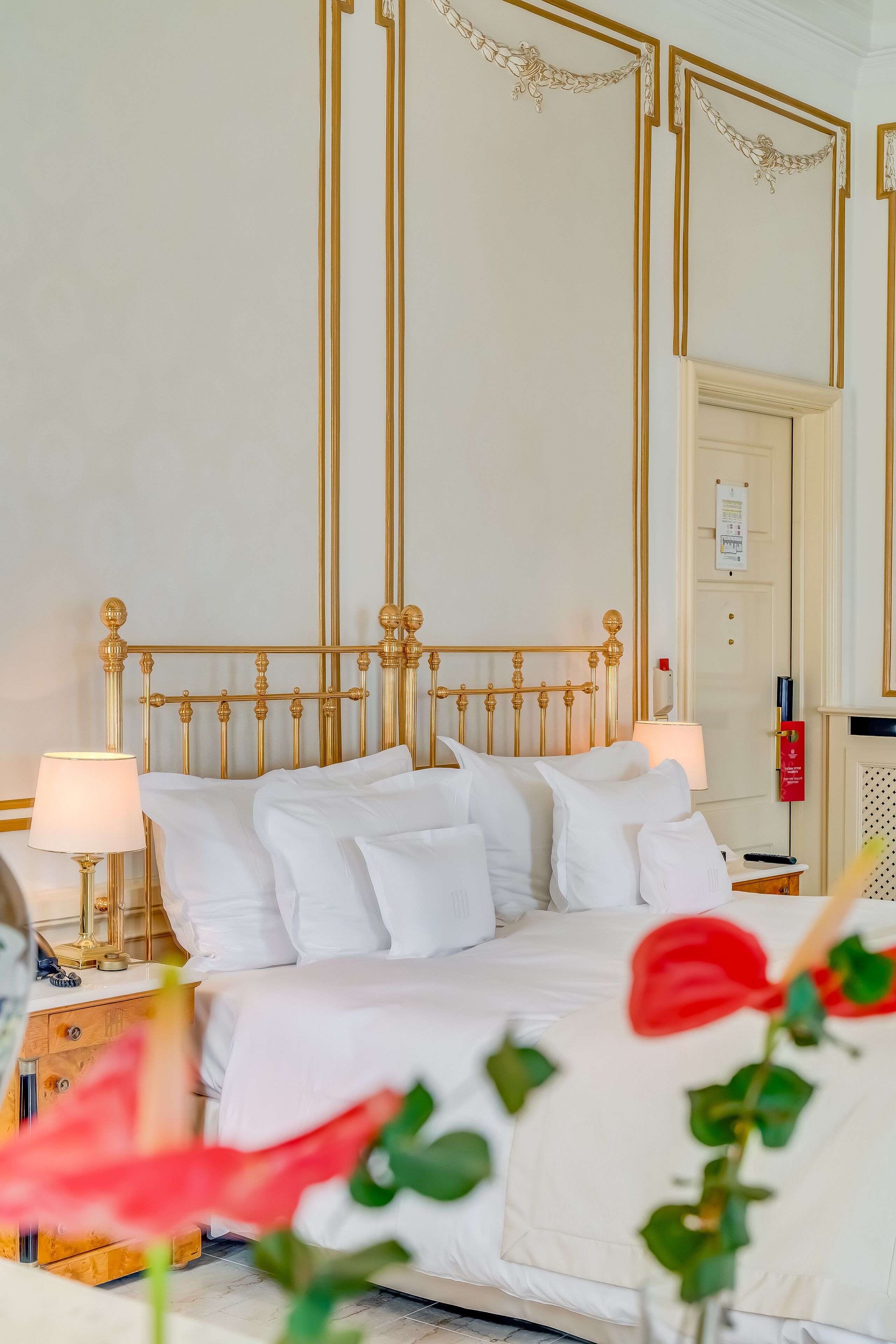 A hotel room with a gold-framed bed, white bedding, and a nightstand with a lamp.