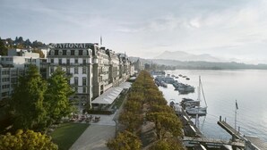A waterfront hotel with a row of buildings, a dock with boats, and a view of the water and mountains in the distance. - Grand Hotel National (Lucerne)