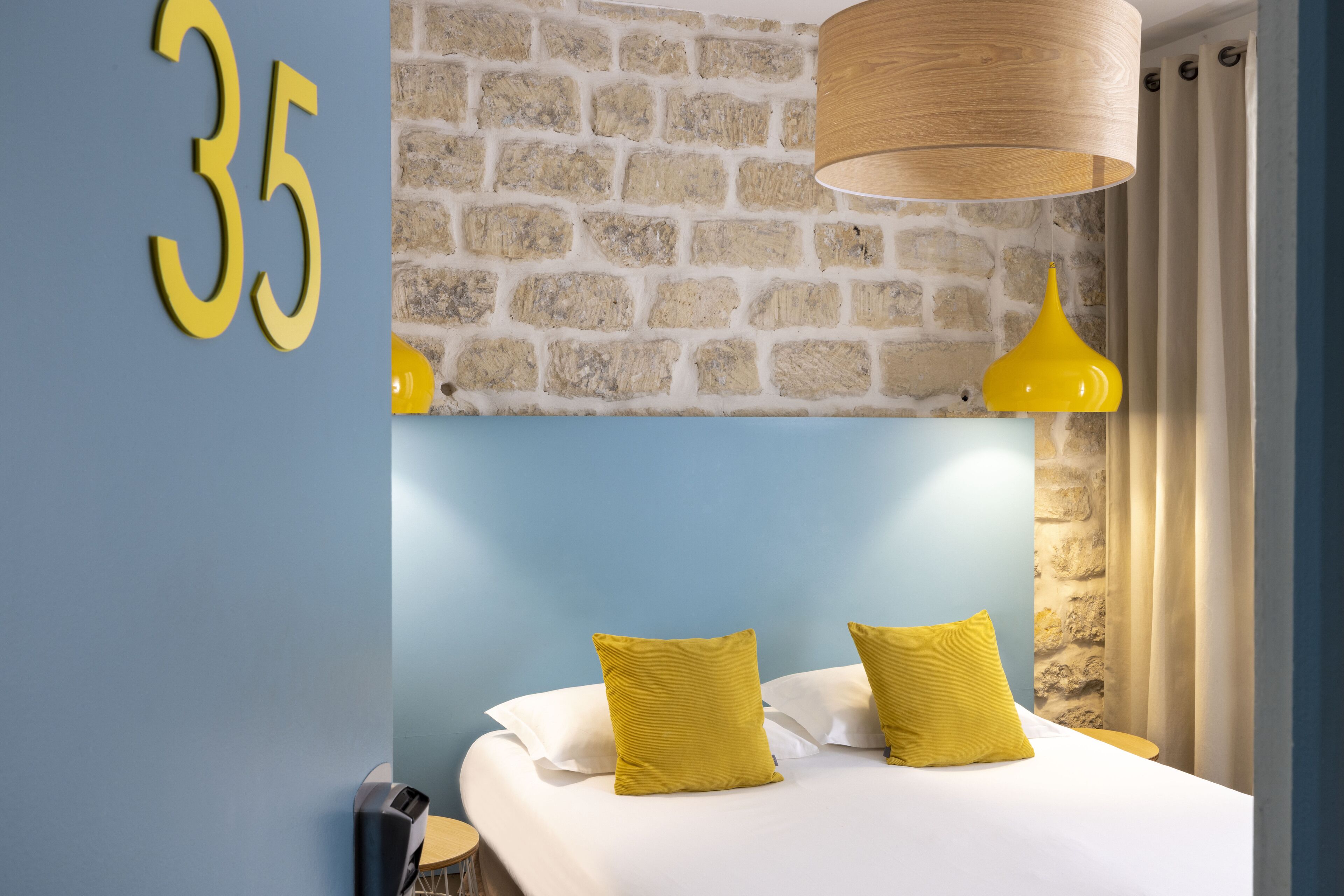 A hotel room with a stone wall, a number '35' sign, a bed with white linens and yellow pillows, and a hanging wooden lamp.