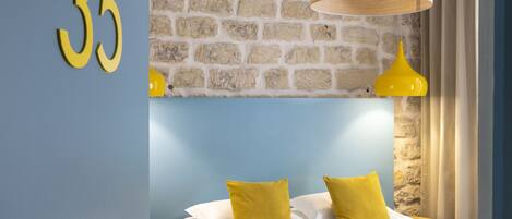 A hotel room with a stone wall, a number '35' sign, a bed with white linens and yellow pillows, and a hanging wooden lamp.