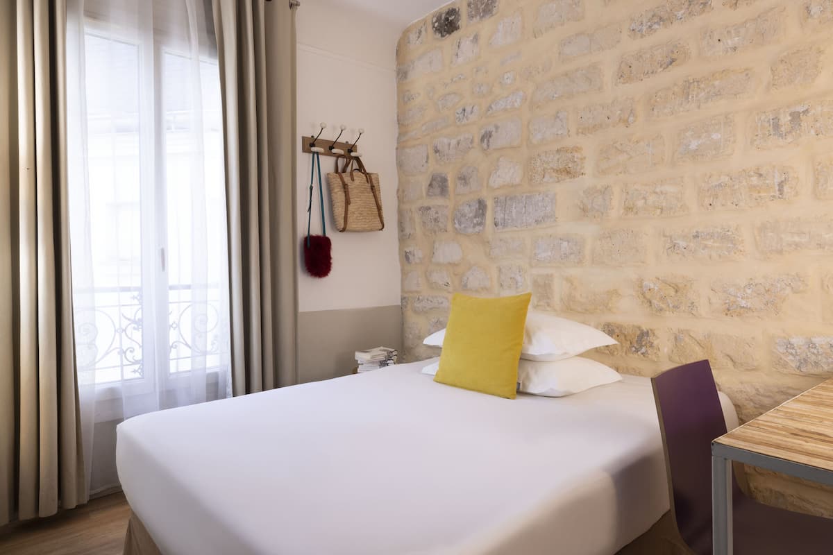 Een hotelkamer met een bed, een houten tafel, een stenen muur en een raam met gordijnen.