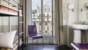A room with a bunk bed, a chair, a bathroom with a sink and mirror, and a balcony with a view of a building.