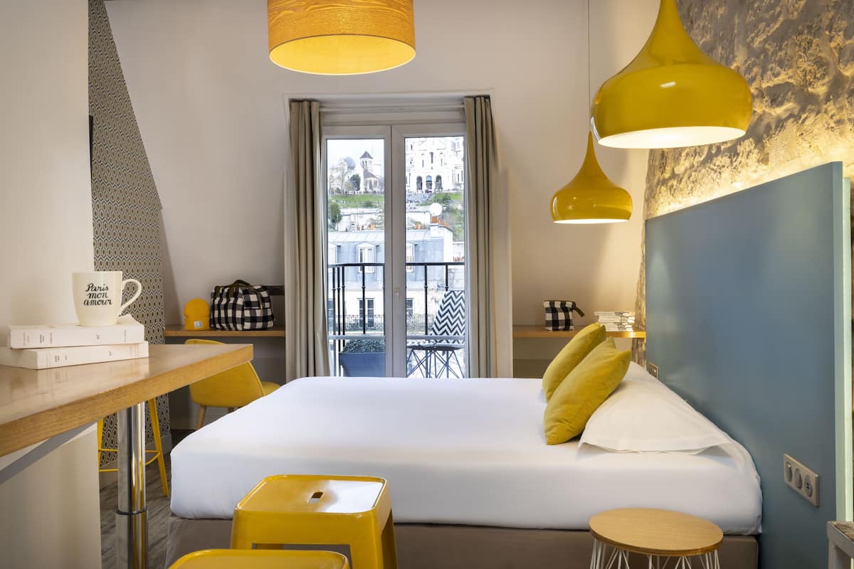 Een hotelkamer met een bed, een bureau met stoel, een klein krukje, een lamp en uitzicht op de stad door het raam.