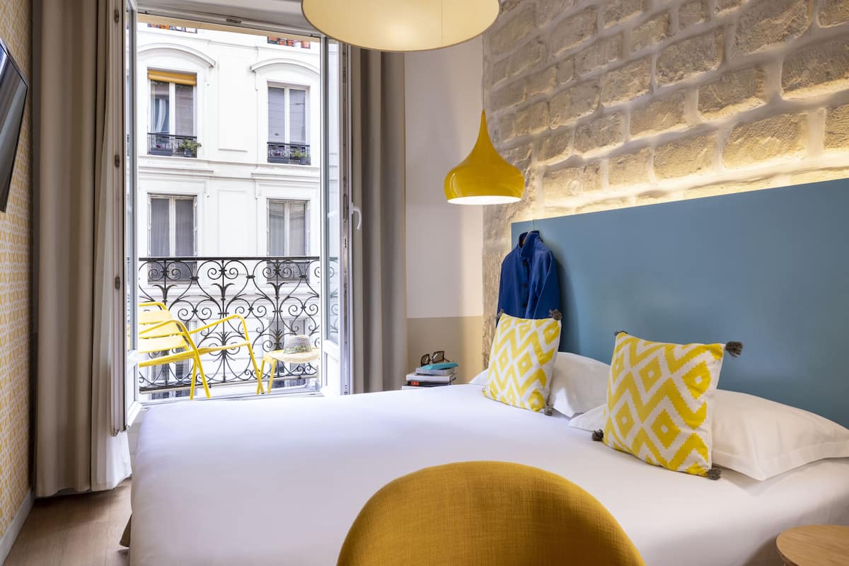 Hotelkamer met een groot bed, uitzicht op een balkon met gele stoelen, een bakstenen muur en een hangende gele pendel.