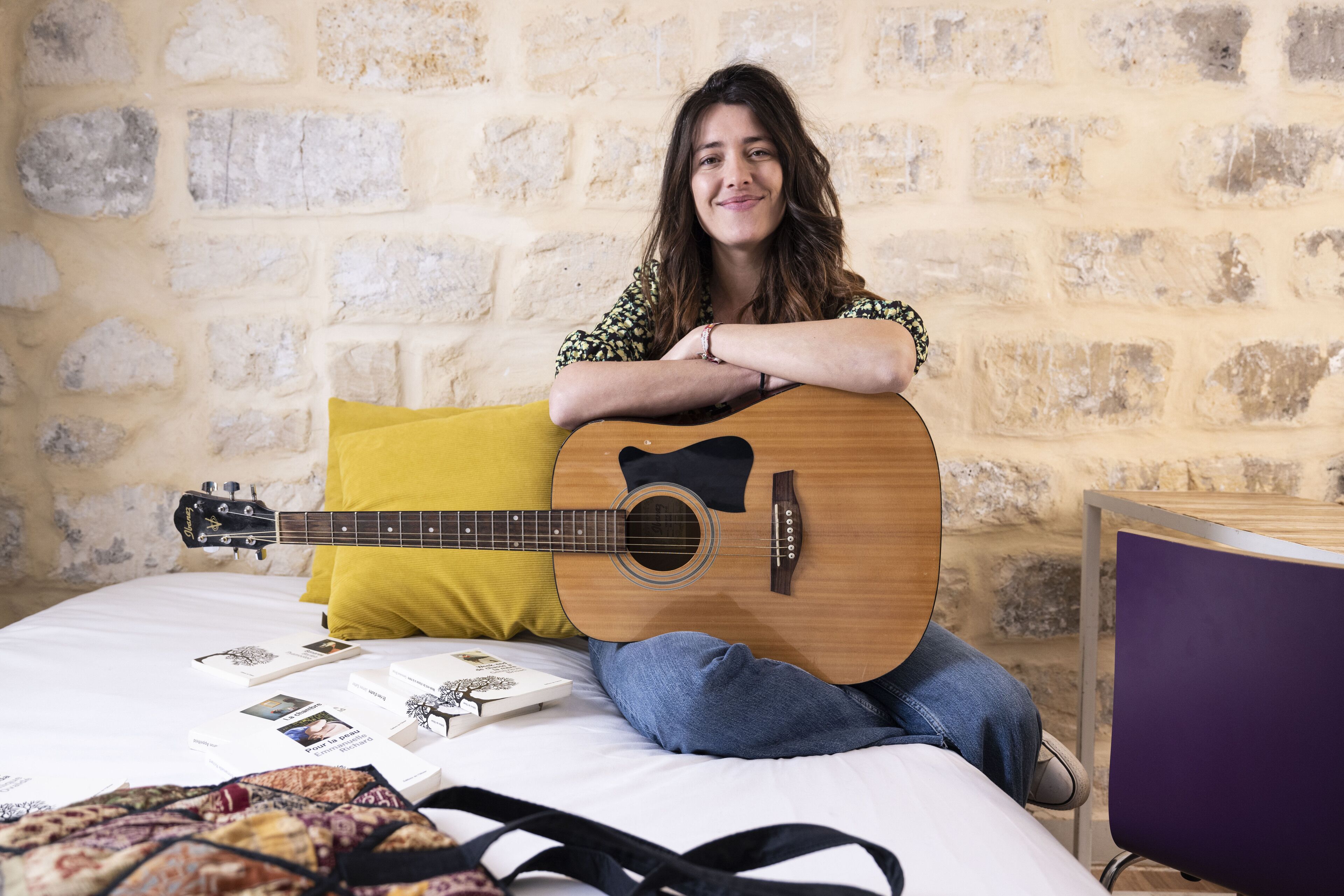 A person sitting on a bed with a guitar, a yellow pillow, and some books.