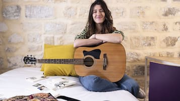 A person sitting on a bed with a guitar, a yellow pillow, and some books.