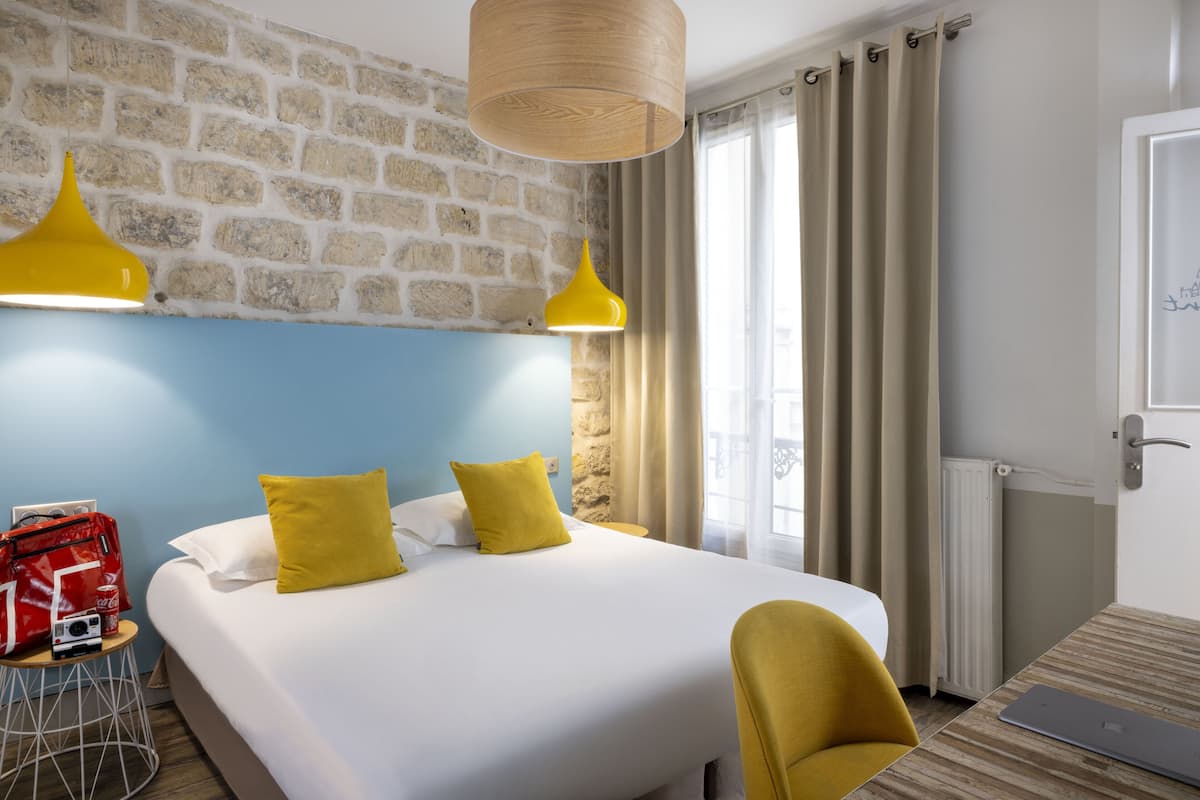 Een hotelkamer met een groot bed, een gele stoel, een rode tas en een laptop op een houten tafel.