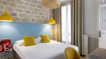 Une chambre d’hôtel avec un grand lit, une chaise jaune, un sac rouge et un ordinateur portable posé sur une table en bois.