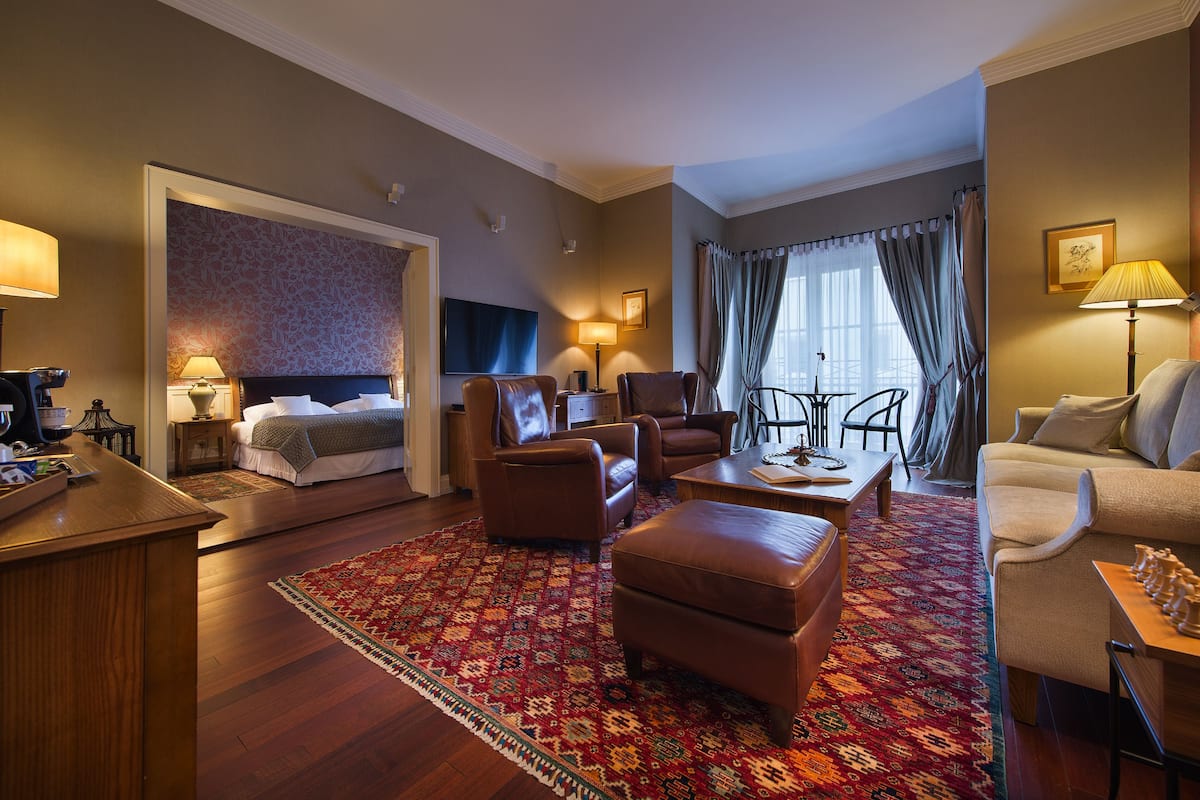Een hotelkamer met een bed, twee fauteuils, een bank, een salontafel, een bureau en een eettafel.