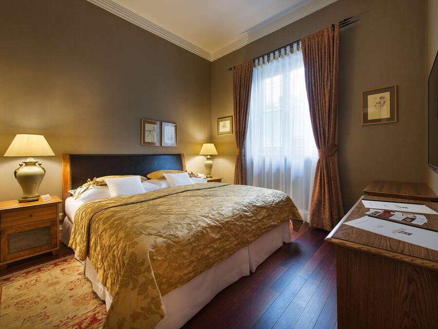 Une chambre d’hôtel avec un grand lit, deux tables de chevet avec des lampes, un bureau en bois et une fenêtre avec des rideaux.