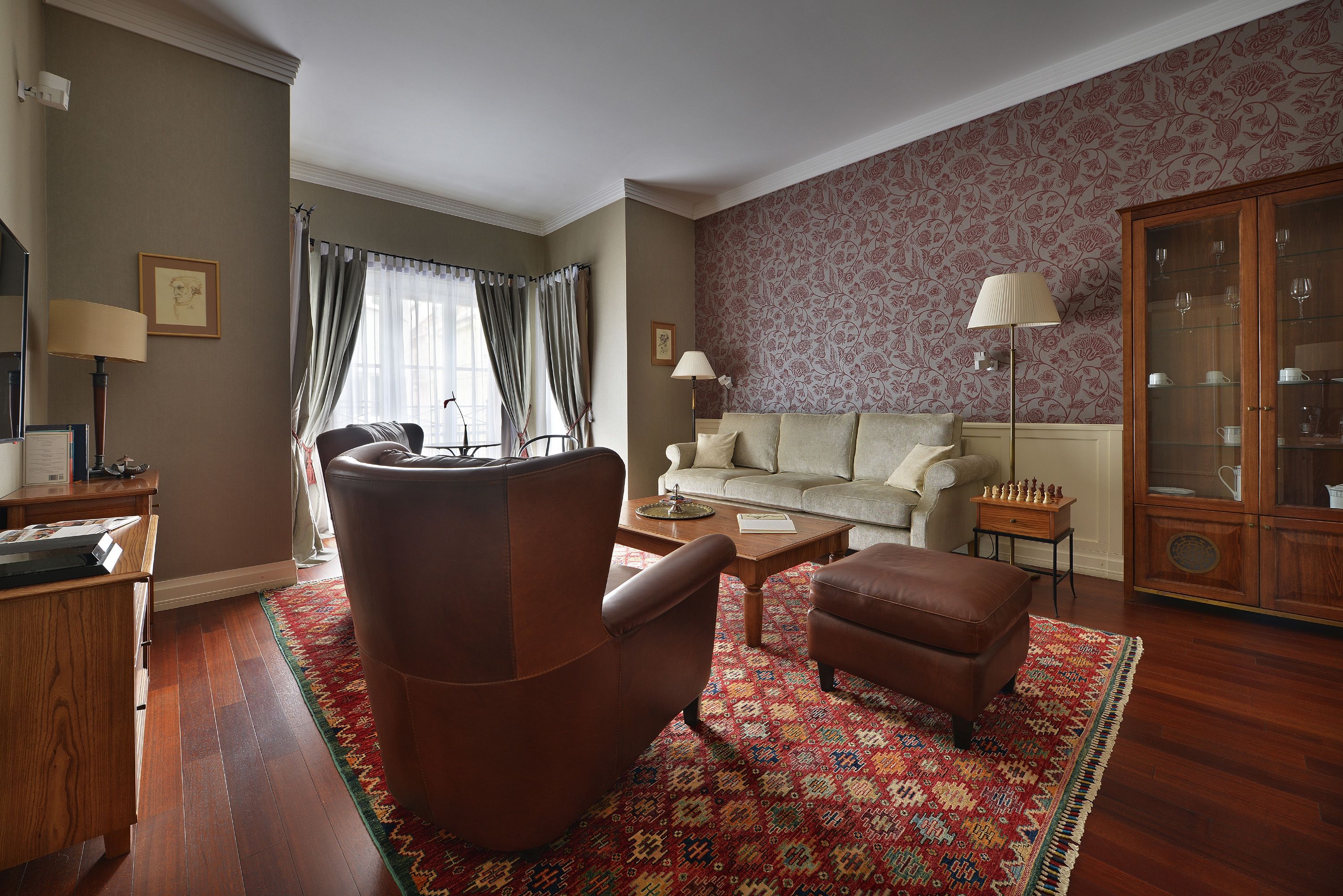 Una sala de estar con un sillón de cuero marrón, un sofá beige, un tapete estampado y un mueble de madera con puertas de cristal.