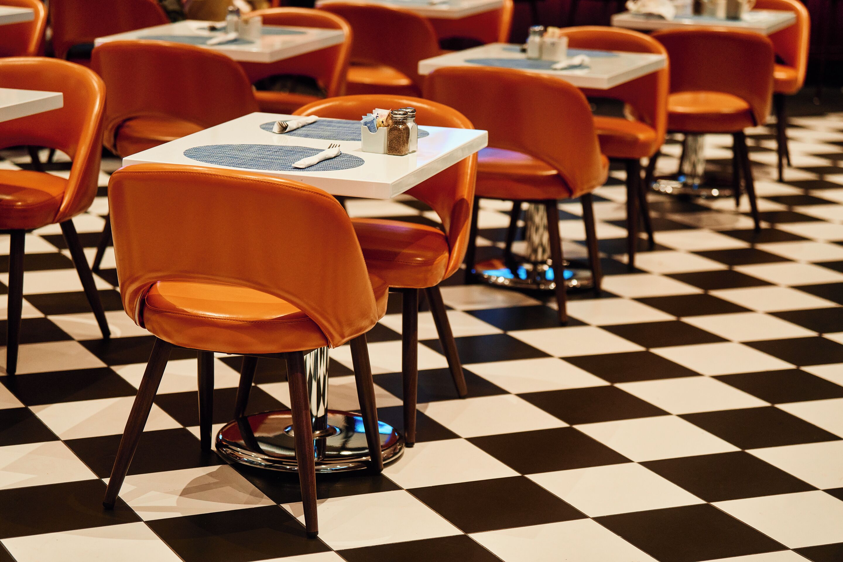Een restaurantinterieur met geruit vloerbedekking, oranje stoelen en witte tafels.