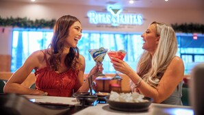 Dos mujeres en un bar dentro de un hotel, con un cartel que dice "Rites & Rituals".