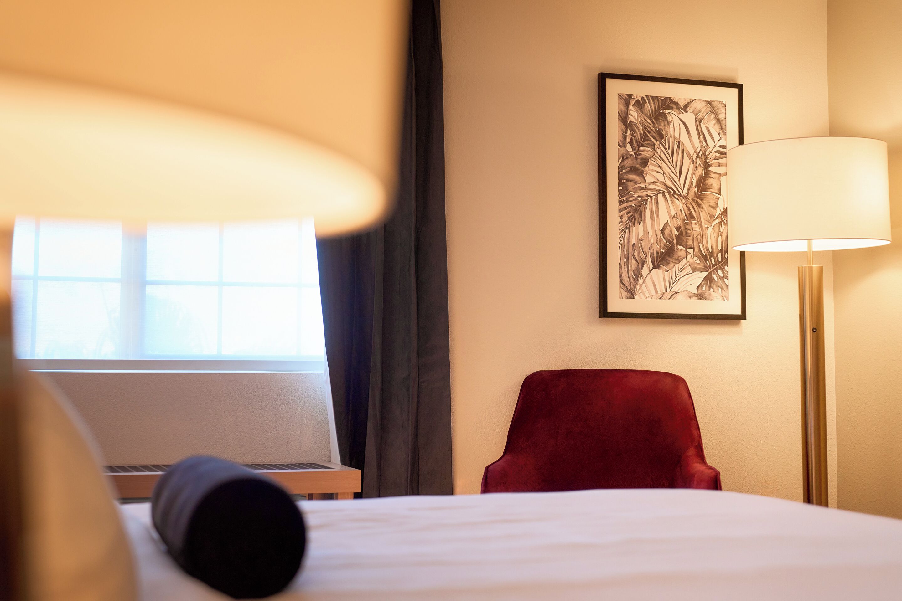 A hotel room with a bed, a desk with a laptop, a red chair, a lamp, and a framed artwork on the wall.