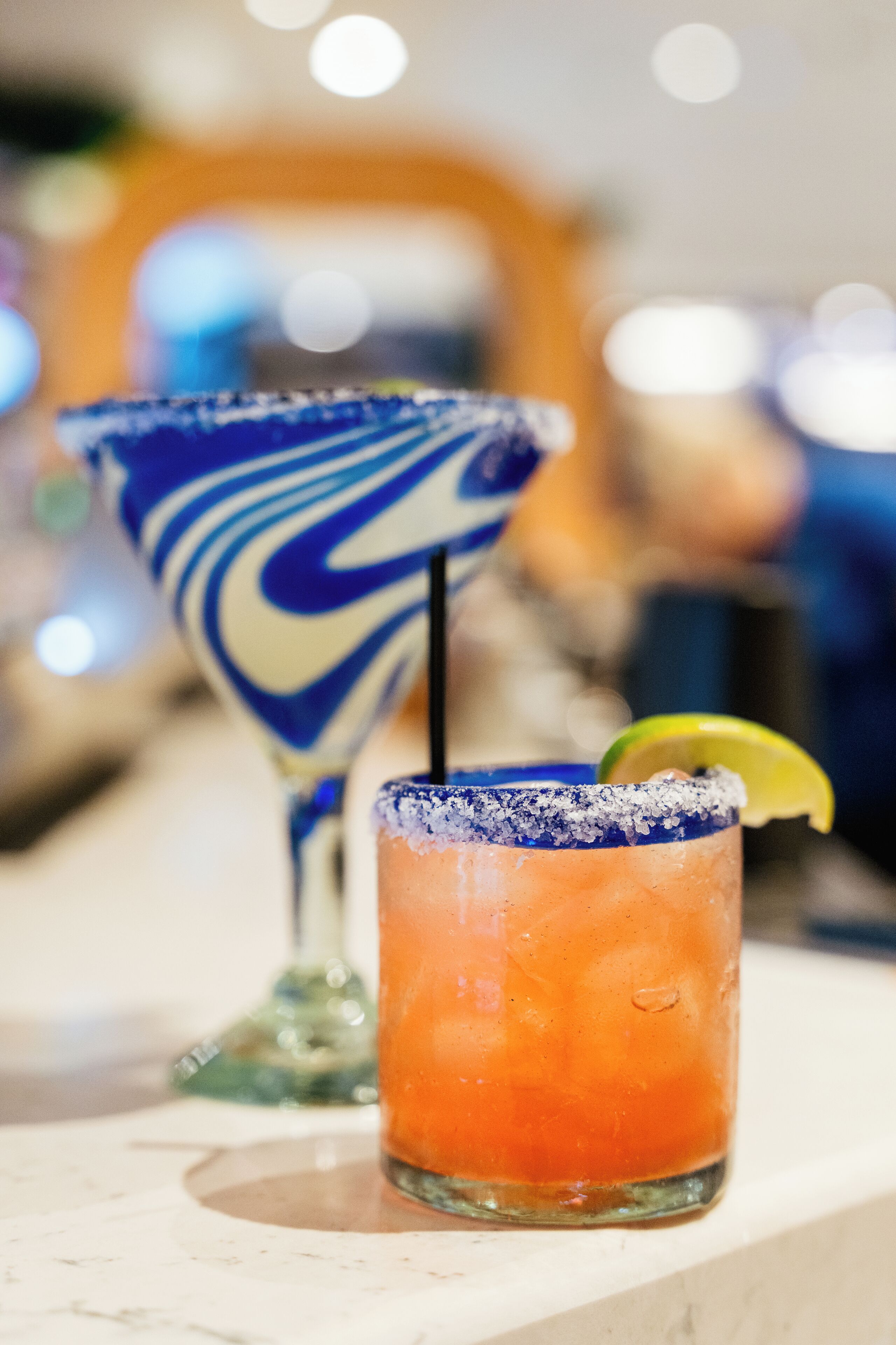 Two cocktails on a bar counter with a blurred background.