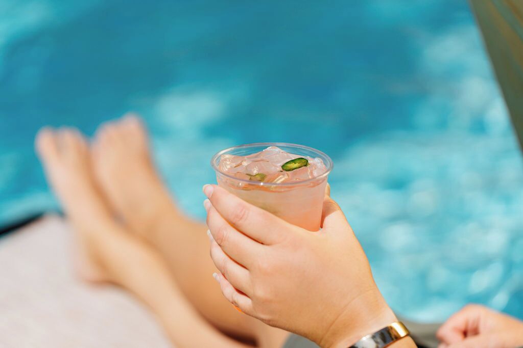 A person holding a drink with a clear cup and ice cubes, with a pool in the background.