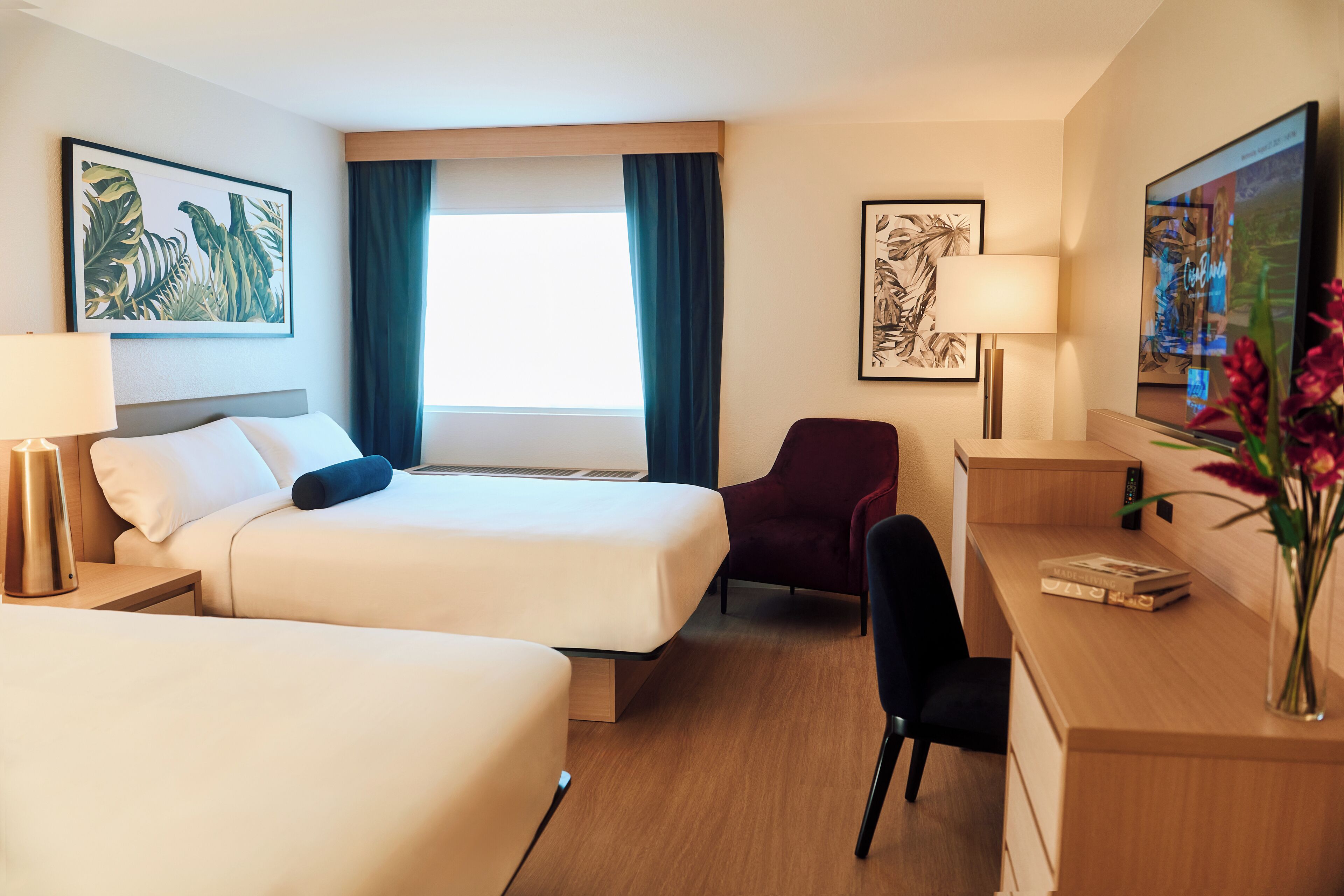 Une chambre d’hôtel avec deux lits, un bureau, une chaise, une lampe et des œuvres d’art accrochées aux murs.