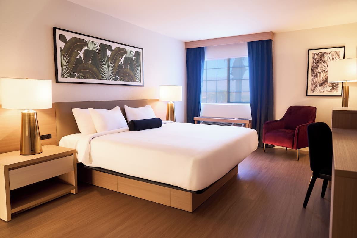 A hotel room with a large bed, two bedside tables with lamps, a red armchair, a desk, and a window with curtains.