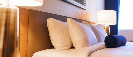 Una habitación de hotel con cama, almohadas, una mesita de noche, una lámpara y un cuadro en la pared.