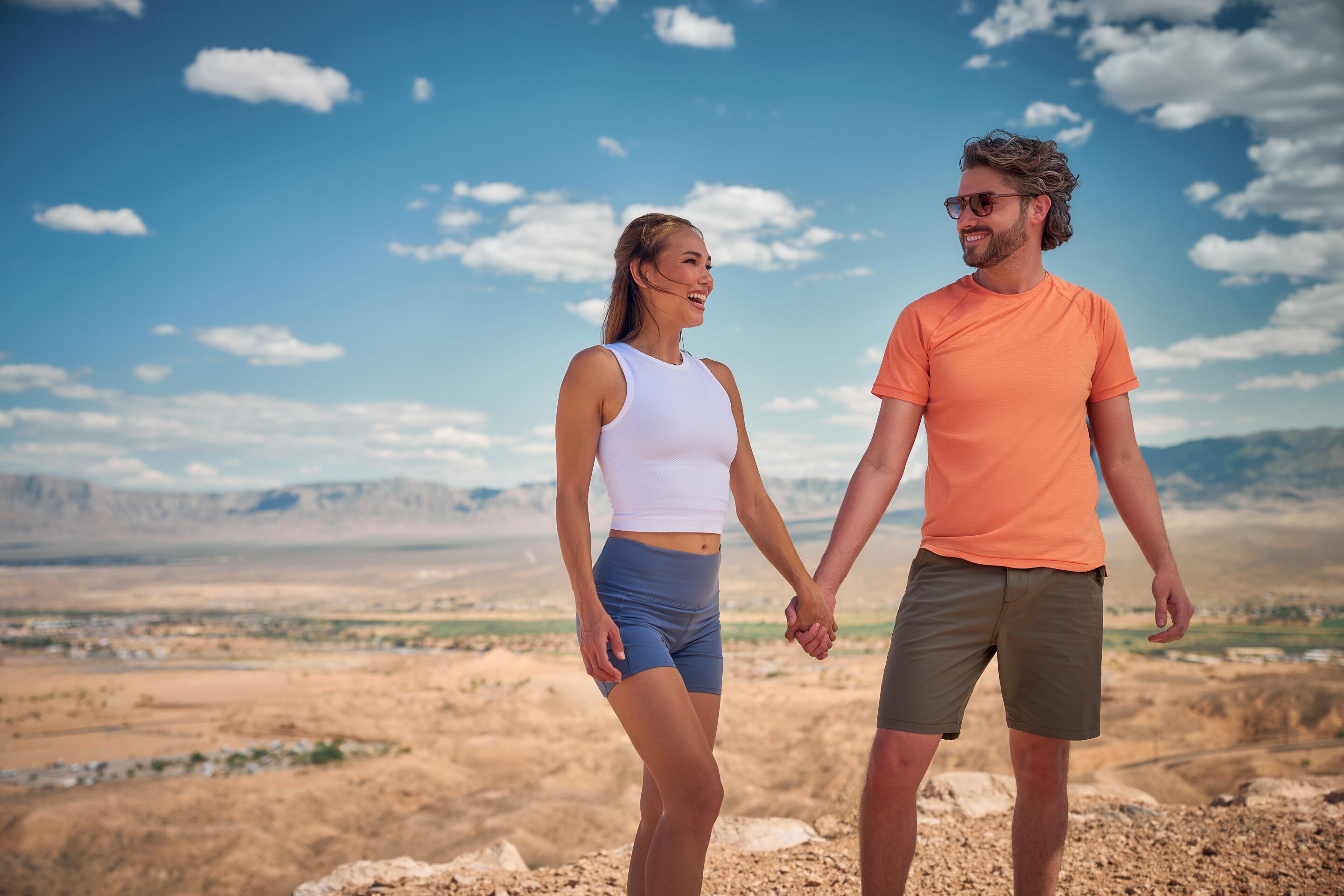 Um casal de mãos dadas em uma paisagem desértica com montanhas ao fundo.