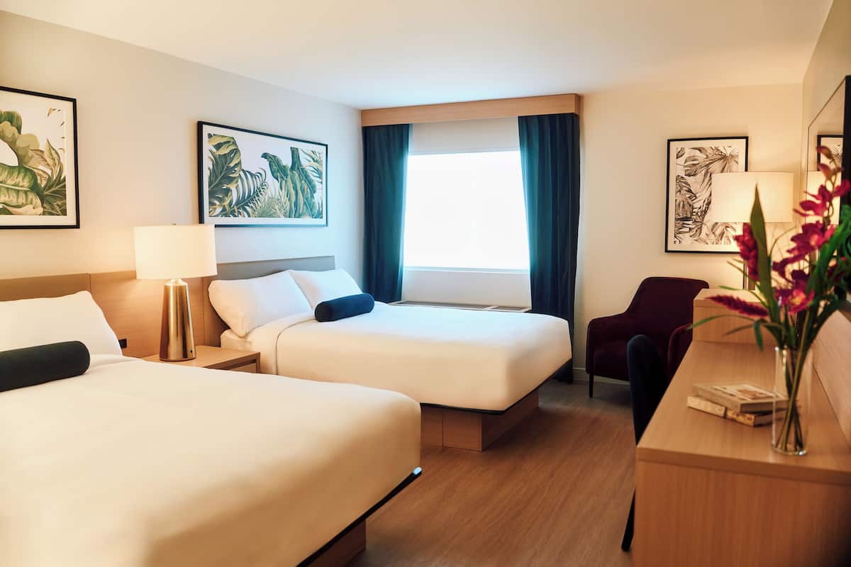 A hotel room with two beds, a desk, a chair, a lamp, and artwork on the walls.