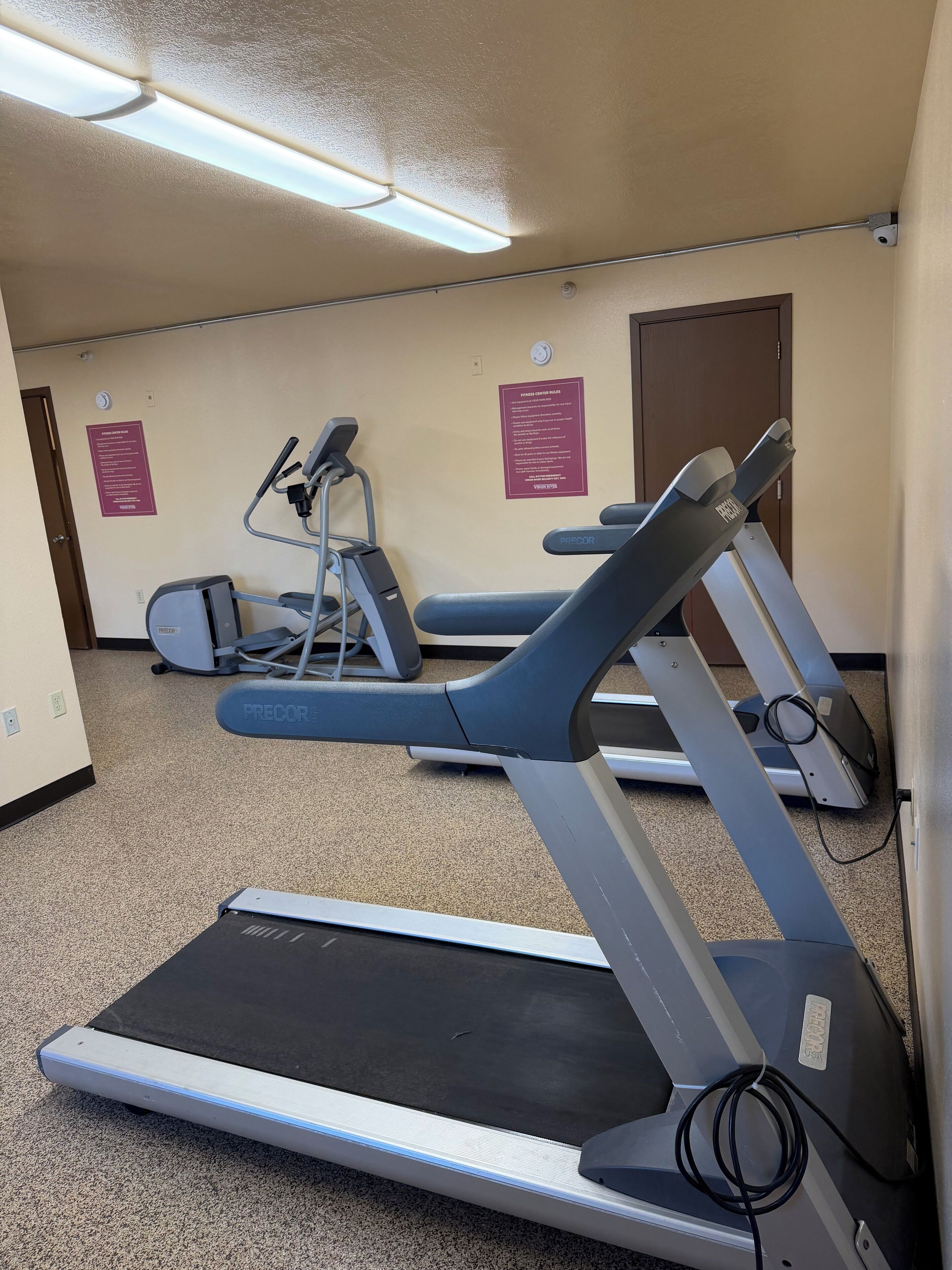 Une salle de sport équipée de tapis de course, d’un vélo d’appartement et d’un appareil d’escalier. On peut voir des panneaux sur le mur et une porte au fond.