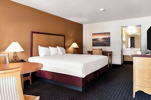 A hotel room with a large bed, two bedside lamps, a desk, a chair, a small table, a mirror, and a painting on the wall.