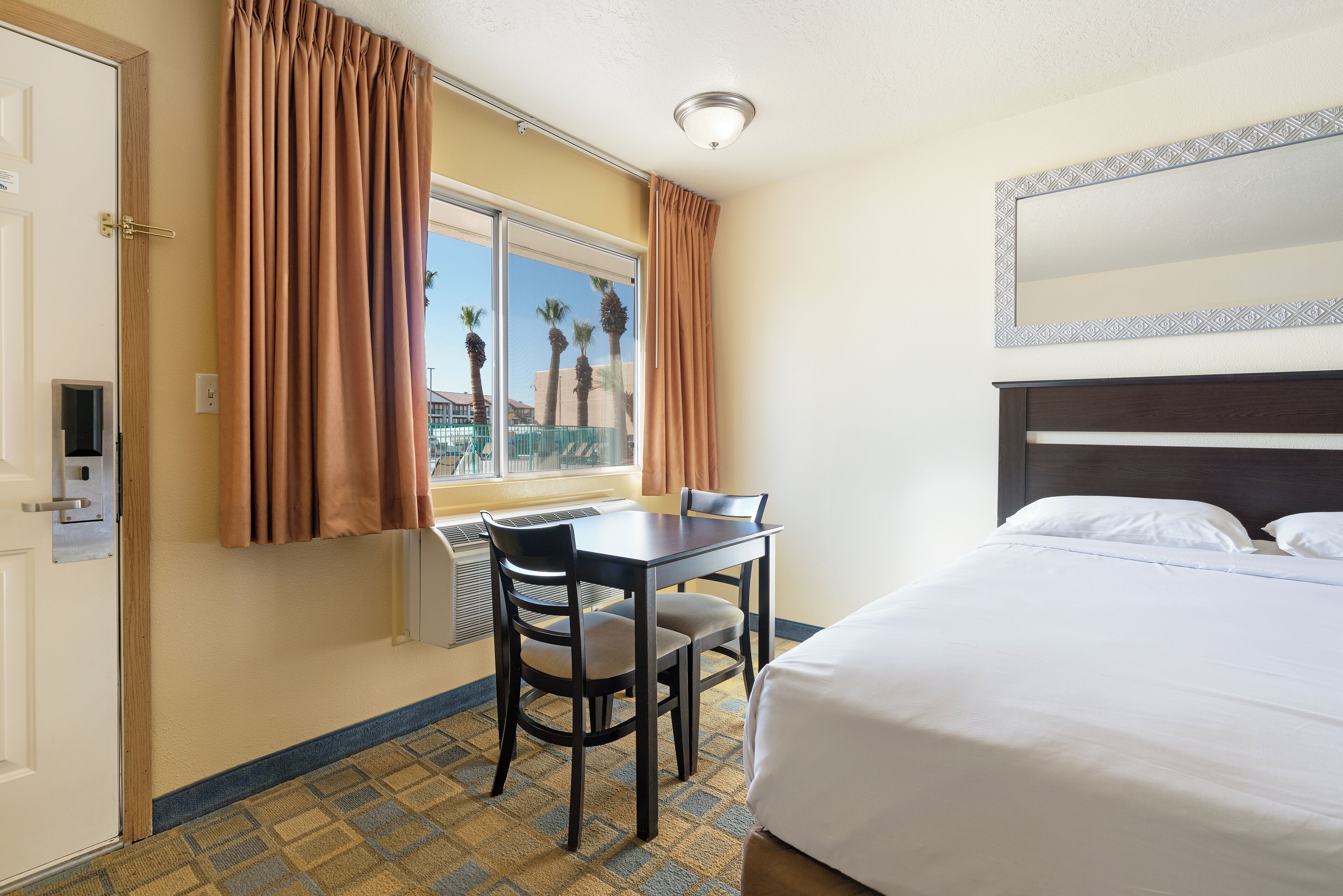 Una habitación de hotel con cama, una mesa y dos sillas, un ventanal con cortinas y vista a palmeras.